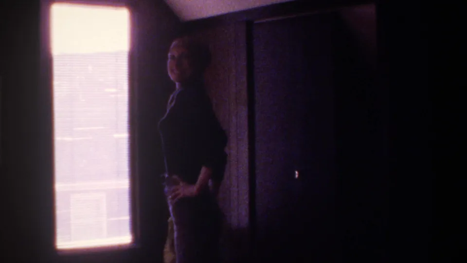8mm interior shot, circa 1975 Florida — woman in dark clothing stands backlit by a large window with horizontal blinds, face partially visible in deep shadow, strong pink-magenta film cast.