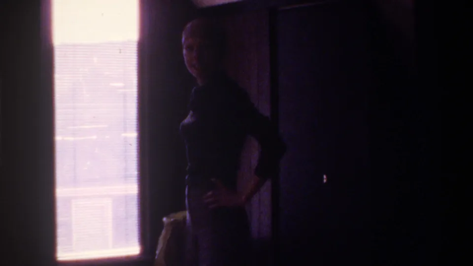 8mm interior, circa 1975 Florida — woman standing near window with horizontal blinds, face slightly more visible than prior frame, heavy pink color cast from aging film emulsion.