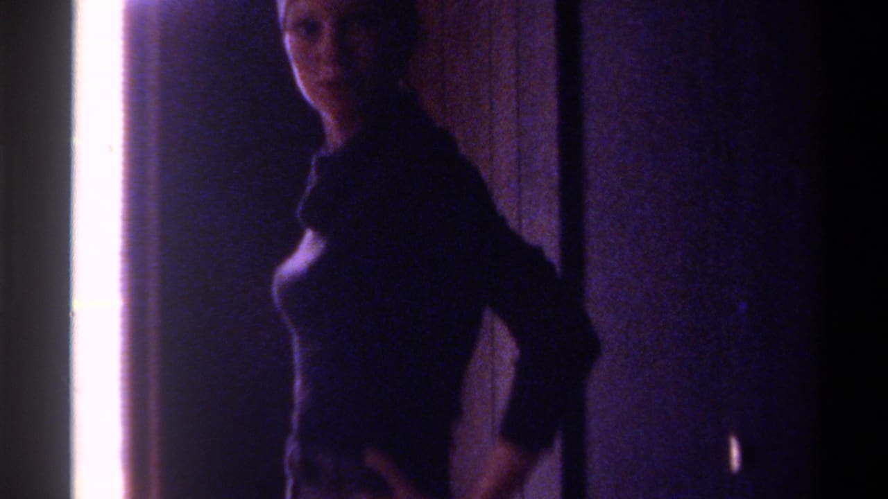 8mm home movie frame, c. 1975, Gainesville, Florida — woman in dark long-sleeve top partially turned toward camera, strong sidelight overexposed at left, dim domestic interior.
