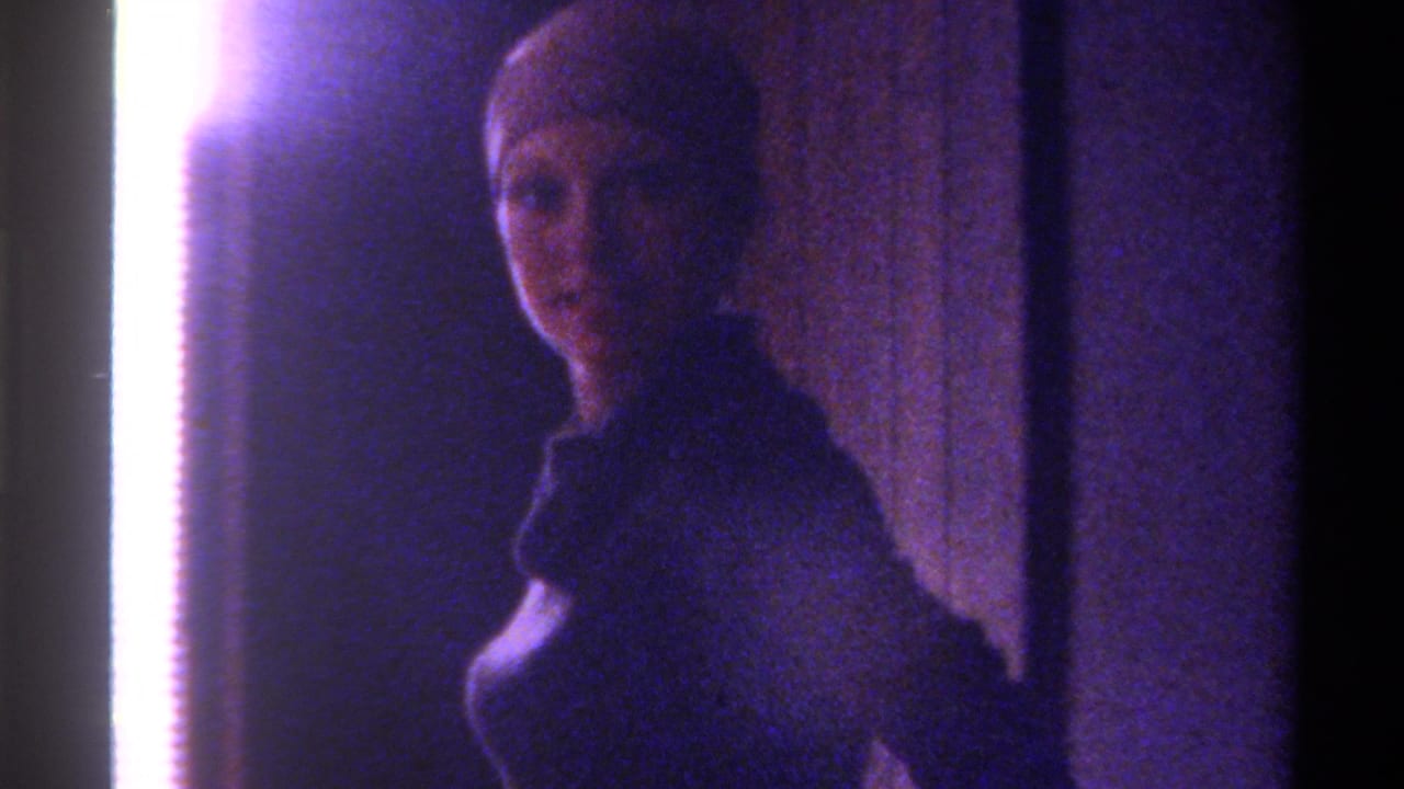 Woman with light hair facing toward camera, dark long-sleeve top, overexposed light strip at left edge, dim interior, cool purple-toned film.