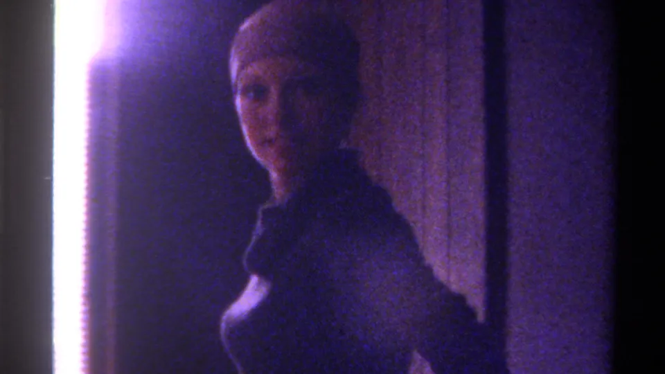 8mm home movie frame, c. 1975, Gainesville, Florida — woman with light hair turning to face camera, strong overexposed sidelight at left, cool blue-purple color cast throughout.