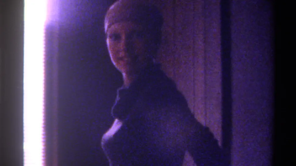 8mm home movie frame, c. 1975, Gainesville, Florida — woman smiling toward camera in low-lit domestic interior, strong overexposed sidelight at left, cool purple film color cast.