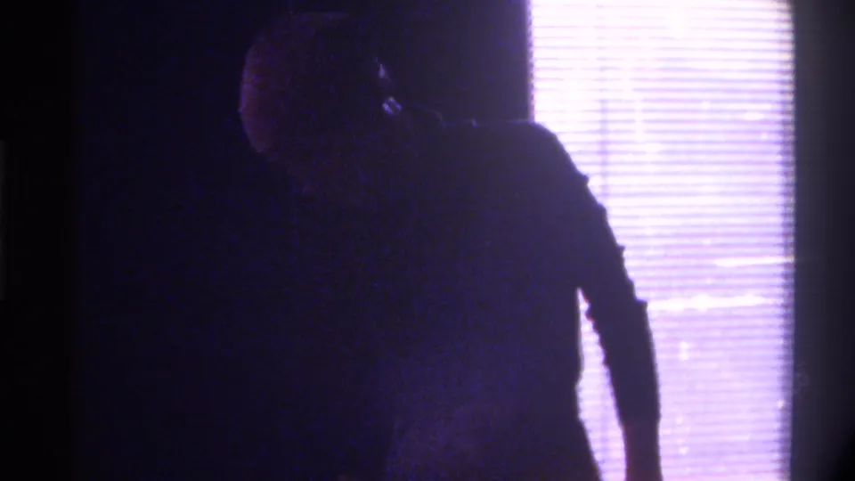 8mm home movie frame, c. 1975, Gainesville, Florida — woman turned away from camera, silhouetted against backlit window with horizontal blinds at right, dim domestic interior.