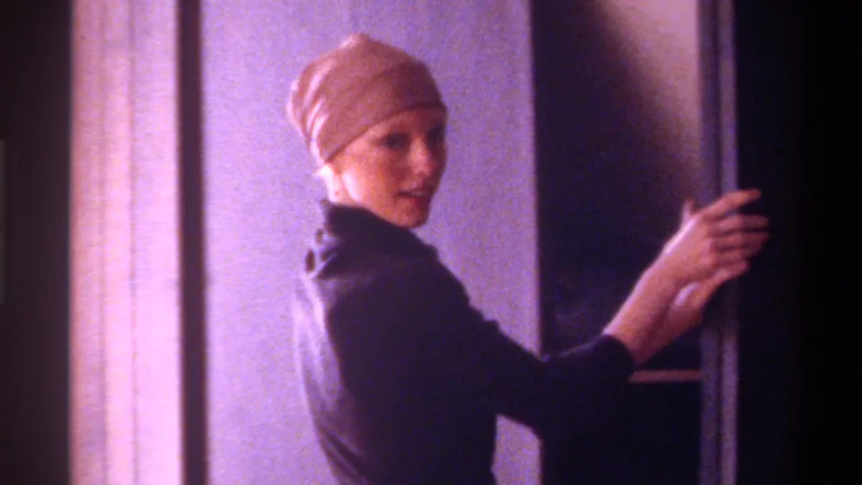 Woman wearing dark turtleneck and beige hair cap, arm extended toward a doorframe, smiling at the camera in a domestic interior, Gainesville Florida circa 1975, 8mm.