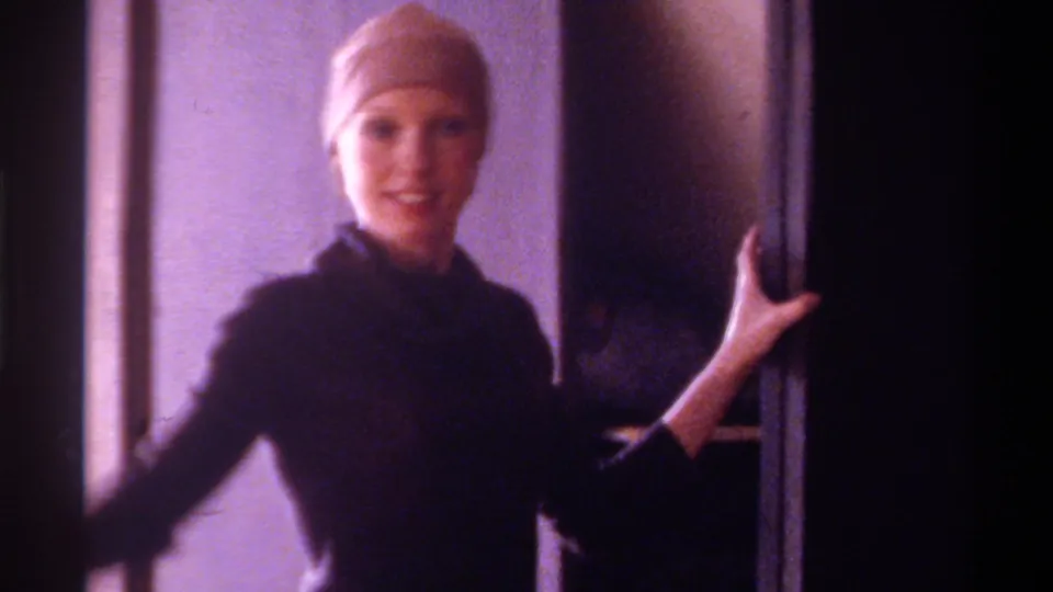 Woman in dark turtleneck and beige hair cap, smiling and posing with arms outstretched near a window in a domestic interior, Gainesville Florida circa 1975, 8mm.