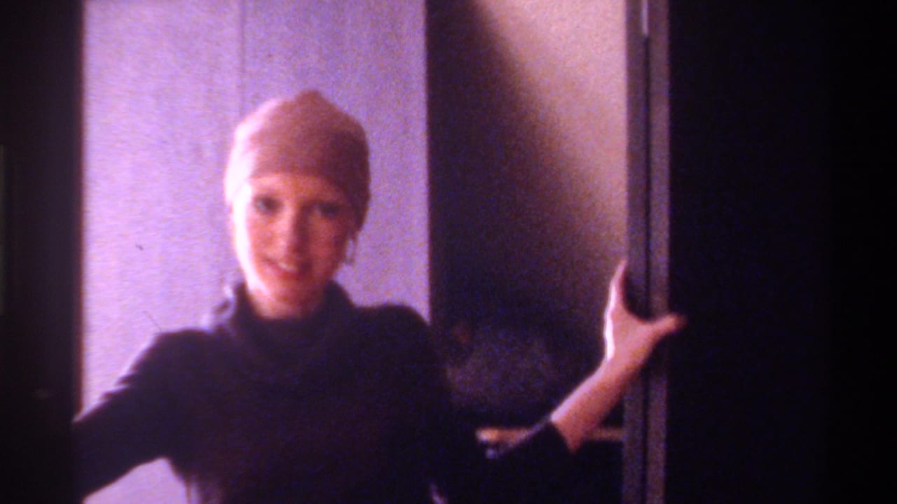 Young woman in black turtleneck and brown head wrap smiling at camera, hand resting on a dark door frame, indoors.