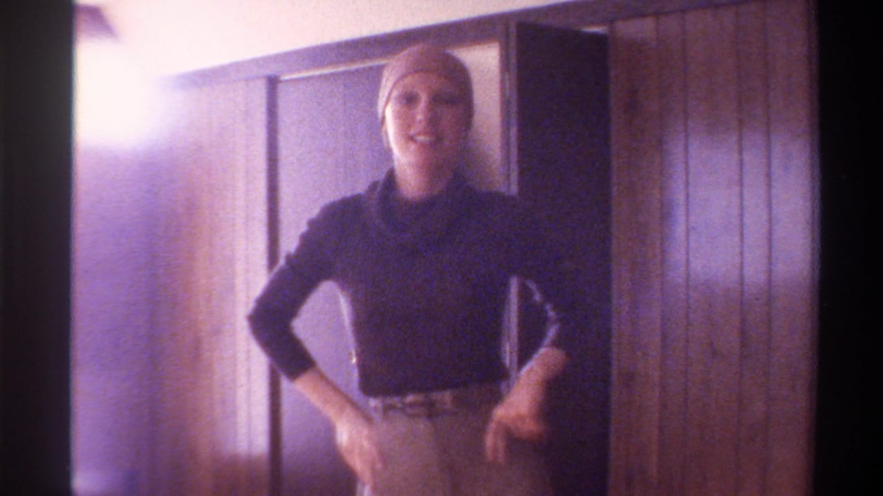 Woman in navy turtleneck and light head wrap stands smiling in a wood-paneled domestic interior; Gainesville, Florida, c. 1975, 8mm home movie.