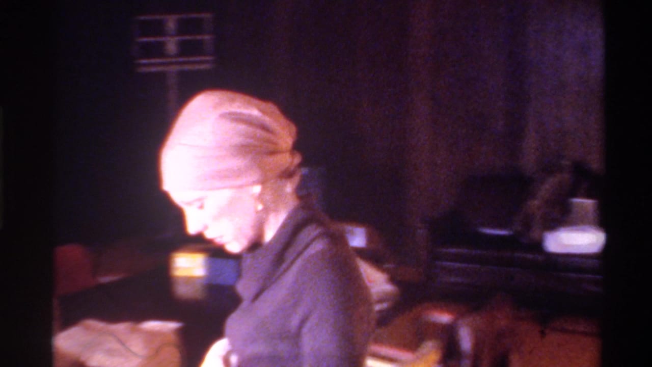 Close profile of woman in light head wrap in a dimly lit wood-paneled room with background items including colored boxes; c. 1975, 8mm home movie.