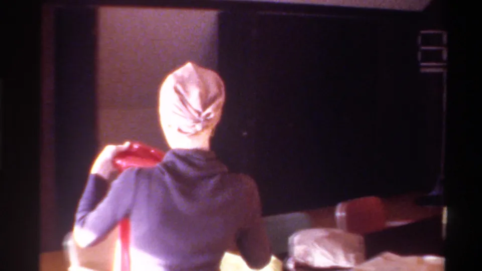 Rear view of woman holding a red bag over a table with wrapped packages in a wood-paneled room; Gainesville, Florida, c. 1975, 8mm home movie.