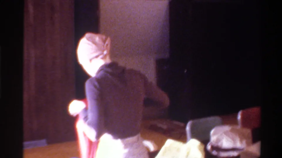 Rear view of woman in dark turtleneck and head wrap at a table with red and pale items in a dim wood-paneled room; c. 1975, 8mm home movie.