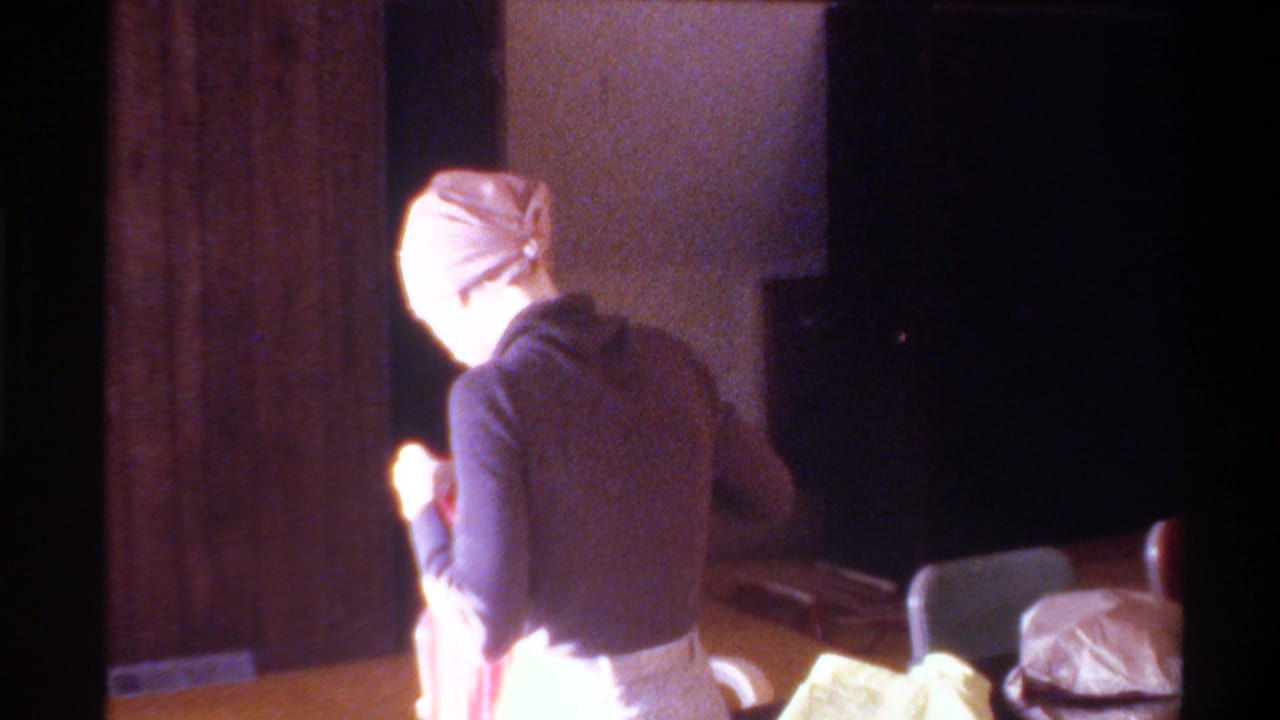 8mm home movie, circa 1975, Gainesville, Florida: woman in dark turtleneck and beige fitted cap, back to camera, beside a table in a domestic interior.