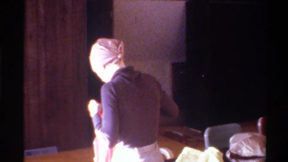 8mm home movie, circa 1975, Gainesville, Florida: woman in dark turtleneck and beige fitted cap, back to camera, beside a table in a domestic interior.