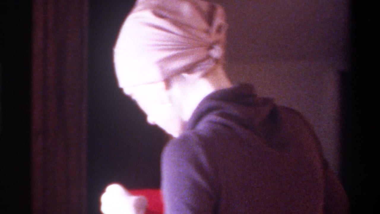 8mm home movie, circa 1975, Gainesville, Florida: woman in beige fitted cap seen in profile, red card visible at frame right, domestic interior with dark background.