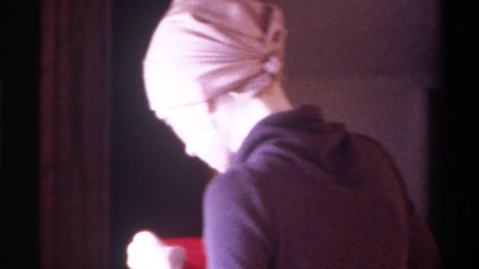 8mm home movie, circa 1975, Gainesville, Florida: woman in beige fitted cap seen in profile, red card visible at frame right, domestic interior with dark background.