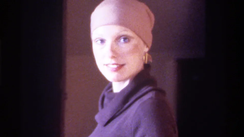 8mm home movie, circa 1975, Gainesville, Florida: woman in beige fitted cap and dark turtleneck turns to smile at the camera, gold drop earrings and blue eyes visible.