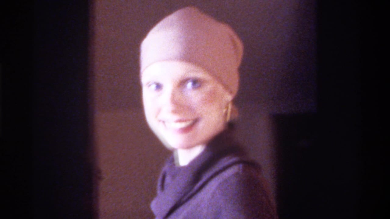 Mid-1970s home movie, Gainesville Florida: woman in pink head wrap and gold drop earring smiles warmly at camera, dark turtleneck, soft indoor lighting.