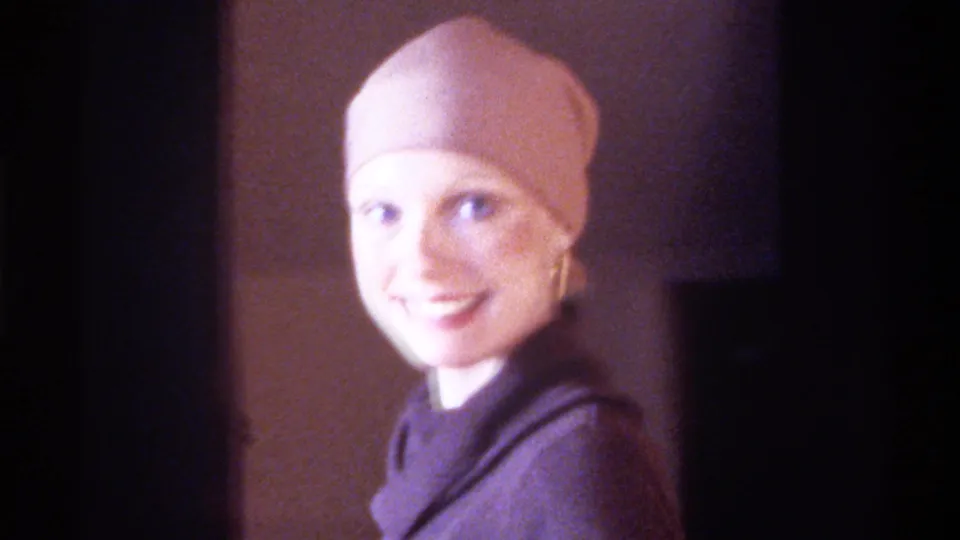 Mid-1970s home movie, Gainesville Florida: woman in pink head wrap and gold drop earring smiles warmly at camera, dark turtleneck, soft indoor lighting.