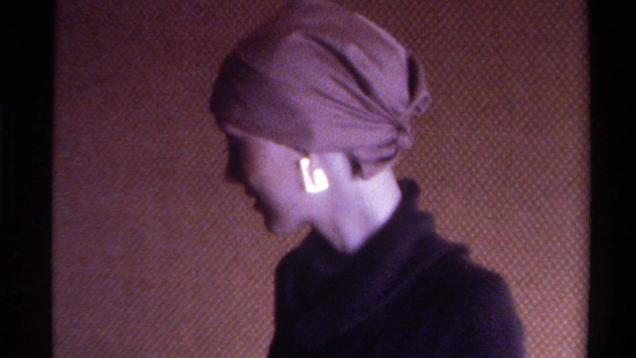Mid-1970s home movie: woman turns to profile revealing head wrap tied at nape and gold drop earring, dark turtleneck, warm brown interior wall.