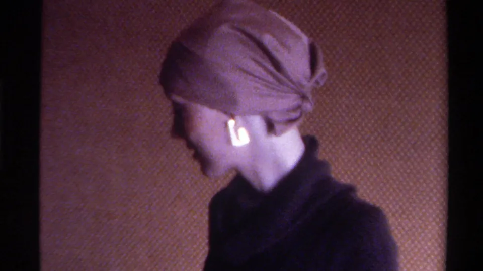 Mid-1970s home movie: woman turns to profile revealing head wrap tied at nape and gold drop earring, dark turtleneck, warm brown interior wall.