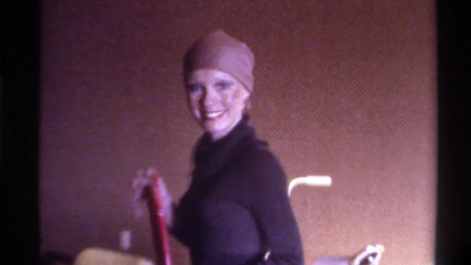 Mid-1970s home movie, Gainesville Florida: woman smiles holding a red-handled object in a domestic interior, head wrap, turtleneck, and partial lamp visible.
