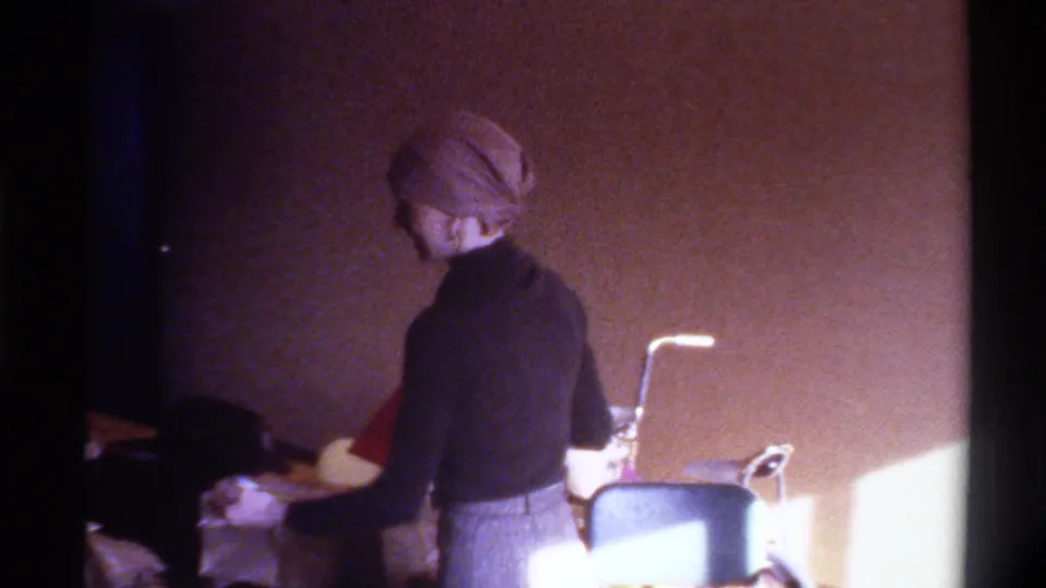 Mid-1970s home movie: woman turned from camera in domestic interior, bent-arm desk lamp and household objects on surface, warm ambient light.