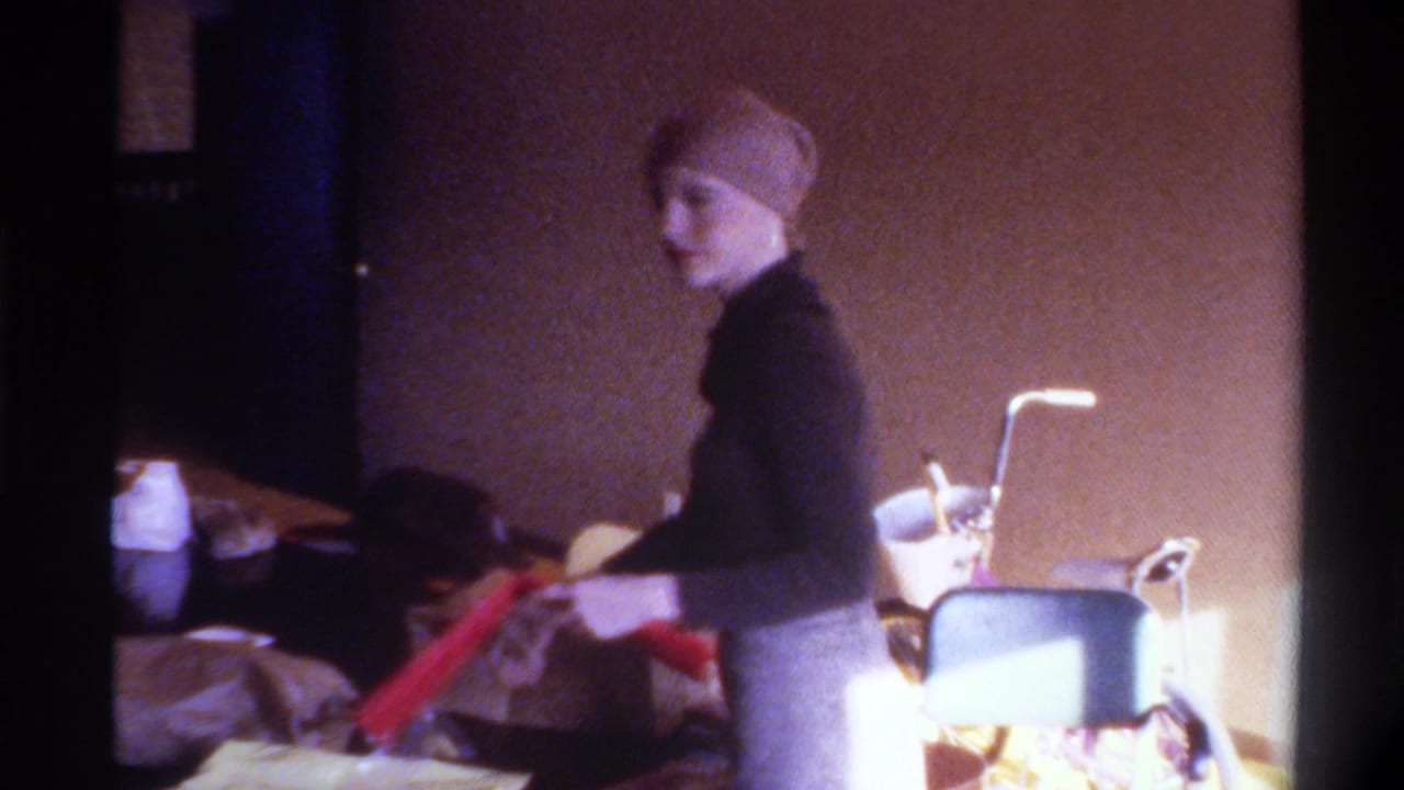 Woman in black turtleneck and tan headscarf stands in profile among wrapped gifts and bags; a wheelchair appears in background of this 1975 Gainesville home interior.