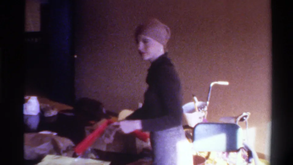Woman in black turtleneck and tan headscarf stands in profile among wrapped gifts and bags; a wheelchair appears in background of this 1975 Gainesville home interior.