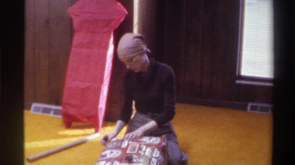 Woman in black turtleneck kneels on gold shag carpet wrapping a patterned gift box; tall red package and venetian blinds visible in this mid-1970s domestic interior.