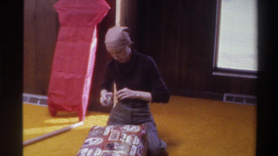 Woman in black turtleneck folds gift wrapping on a patterned box atop gold carpet; tall red package and wood-paneled wall visible in this 1975 home-movie interior.