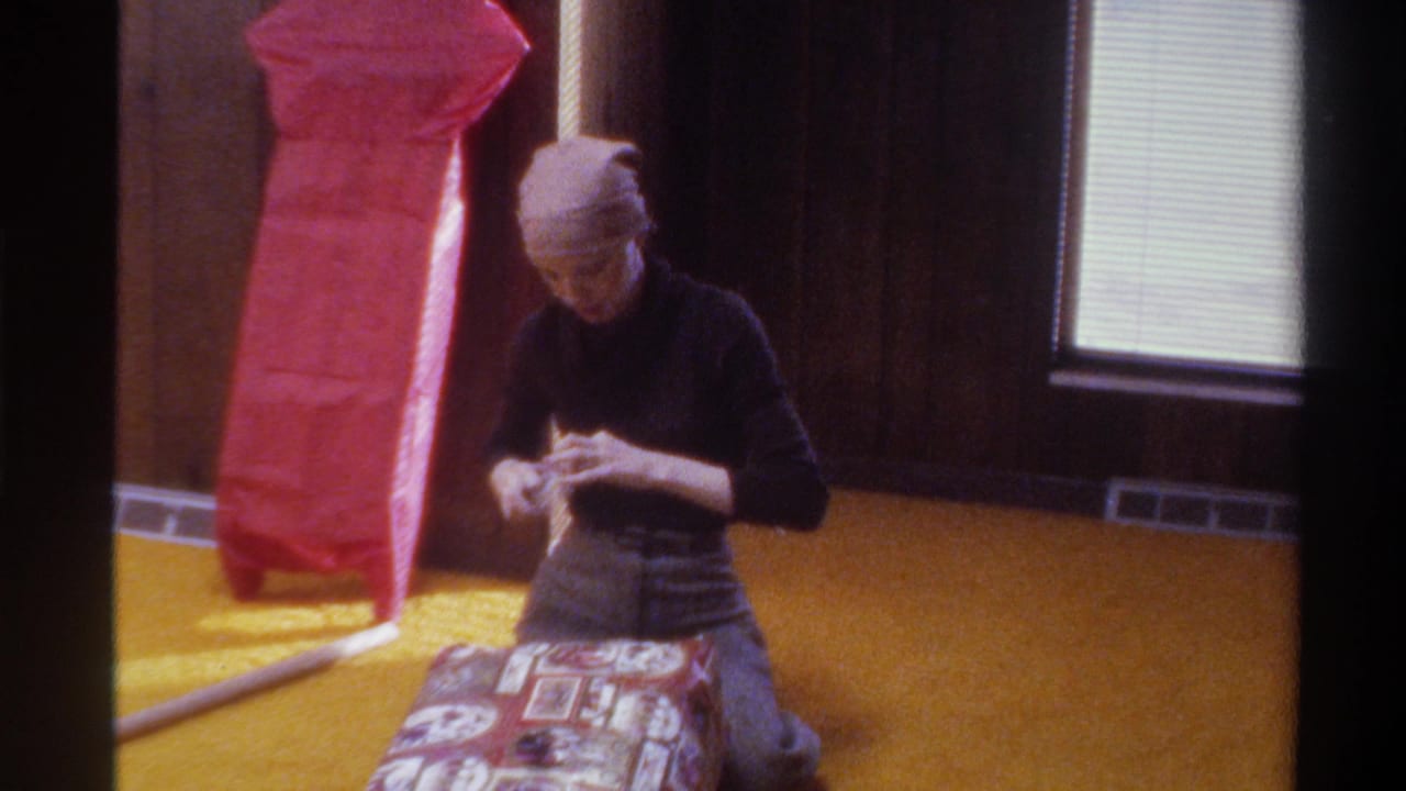 Young woman in 1970s head scarf and dark turtleneck kneels on golden carpet wrapping a patterned gift; wood-paneled walls and red tissue paper visible in background; 8mm home movie, Florida, 1975