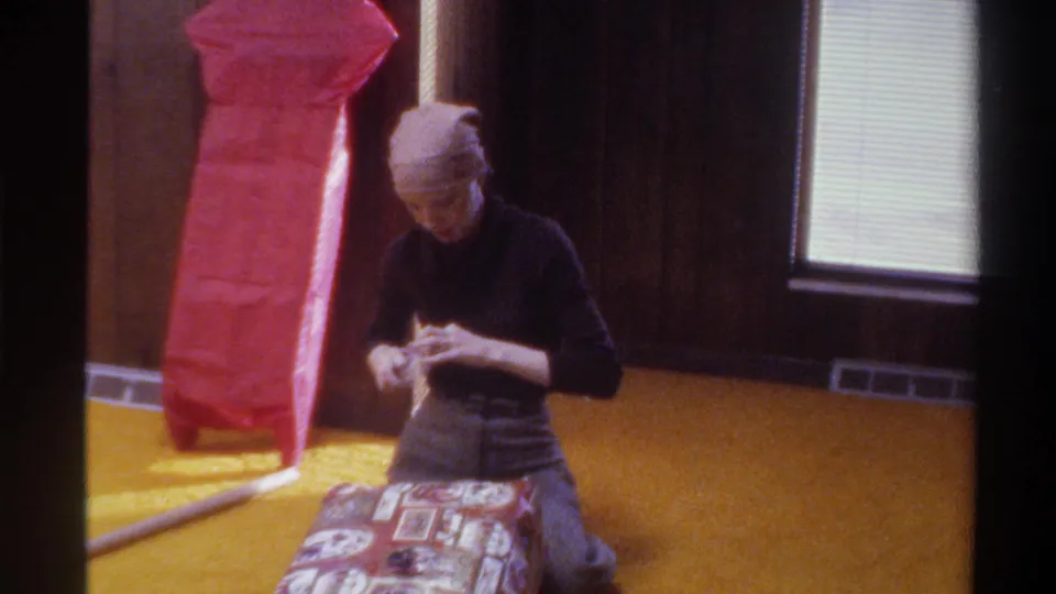 Young woman in 1970s head scarf and dark turtleneck kneels on golden carpet wrapping a patterned gift; wood-paneled walls and red tissue paper visible in background; 8mm home movie, Florida, 1975