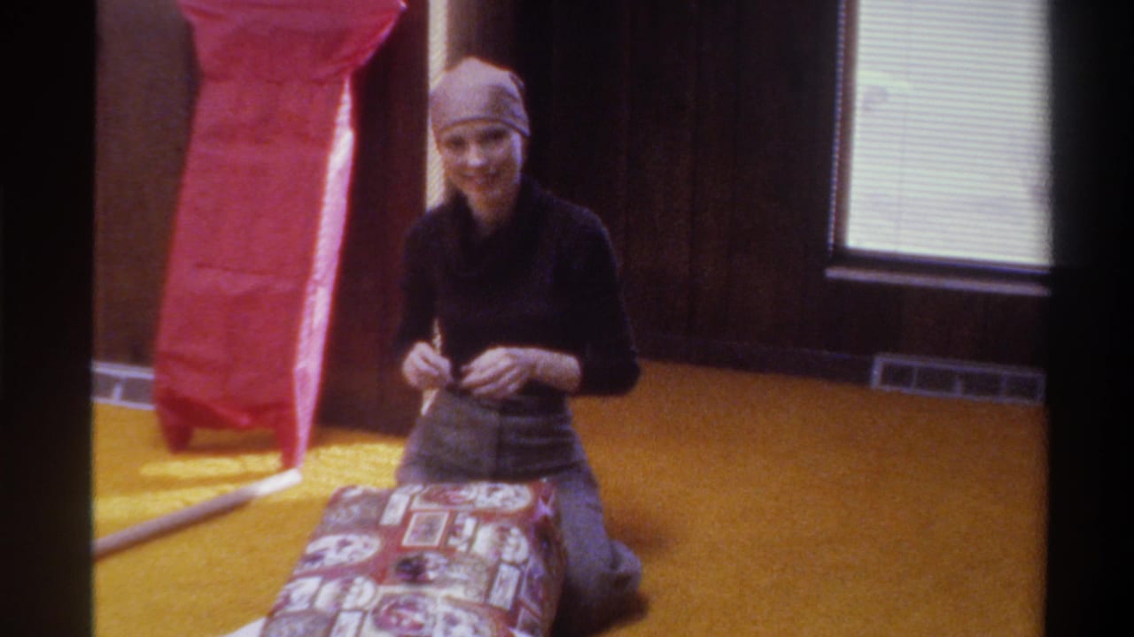 Young woman in 1970s head scarf smiles candidly at the camera mid-gift-wrapping; wood-paneled walls, red tissue paper, and window with blinds behind her; 8mm home movie, Florida, 1975