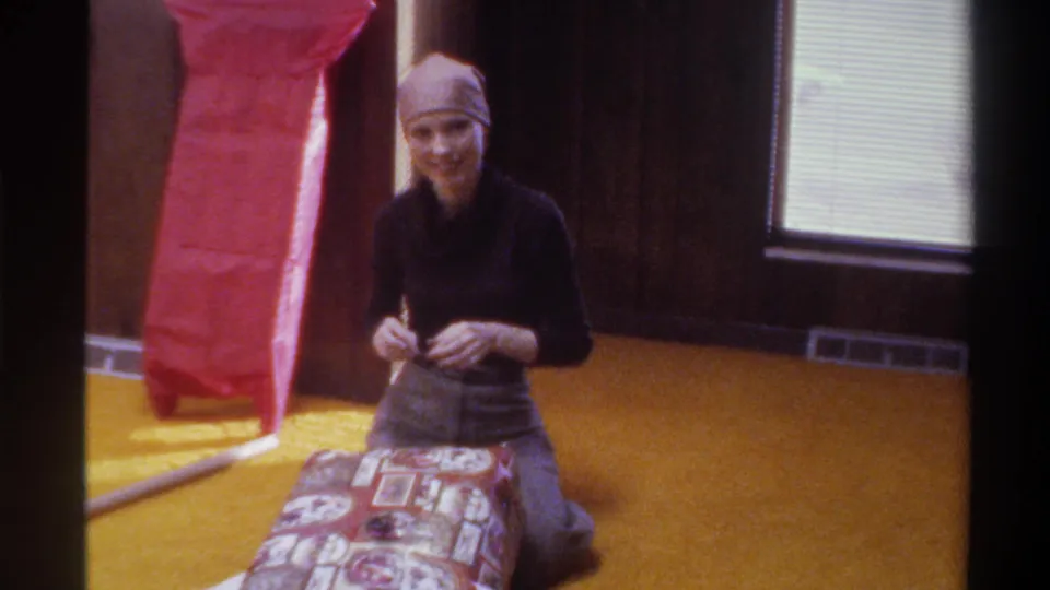 Young woman in 1970s head scarf smiles candidly at the camera mid-gift-wrapping; wood-paneled walls, red tissue paper, and window with blinds behind her; 8mm home movie, Florida, 1975