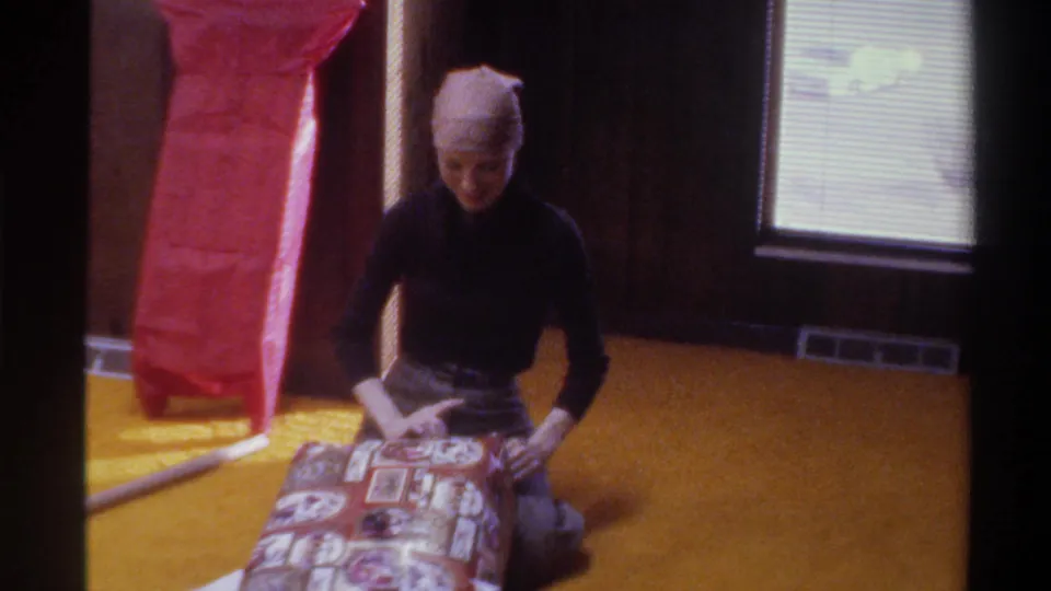 Young woman focused on gift wrapping, bent forward over patterned wrap on golden carpet; baseboard heater, wood-paneled walls, and window with blinds visible; 8mm home movie, Florida, 1975