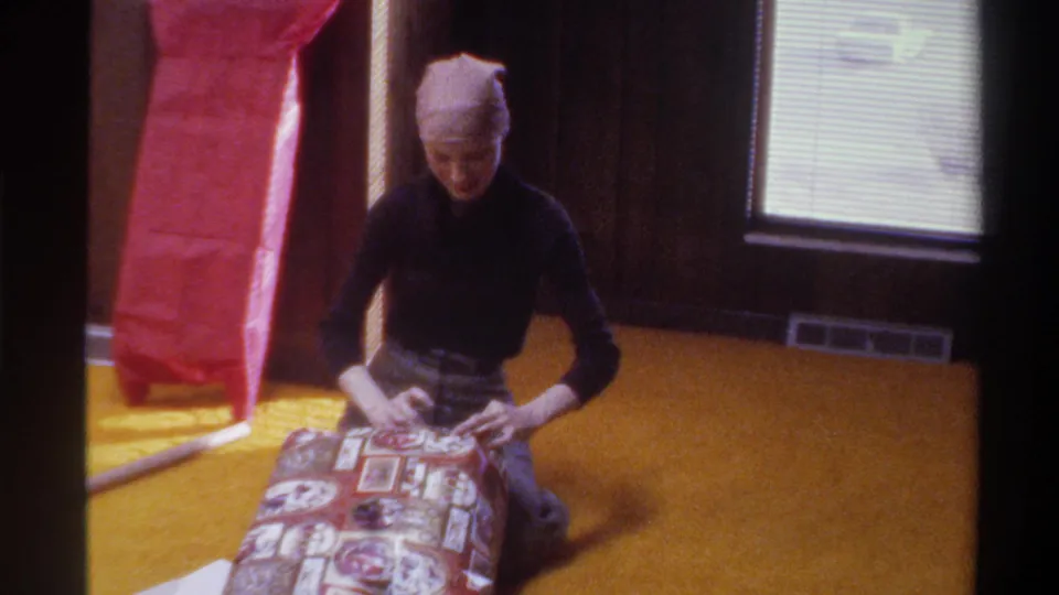 Young woman in 1970s head scarf leans over patterned gift on golden carpet; red tissue paper, dark wood paneling, and window with Venetian blinds in background; 8mm home movie, Florida, 1975