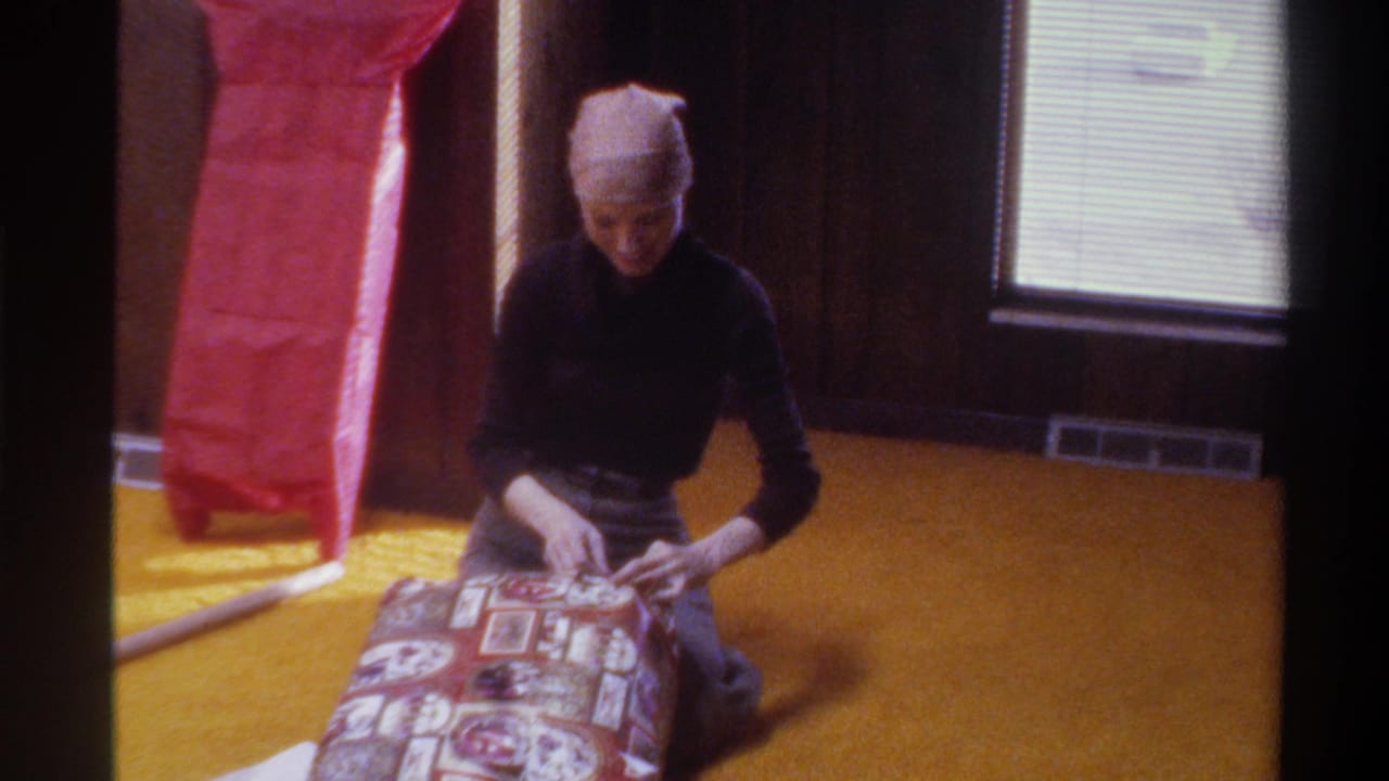 Woman in black turtleneck and knit beanie wraps a gift on yellow carpet in a wood-paneled room, 8mm home movie, Gainesville Florida, circa 1975