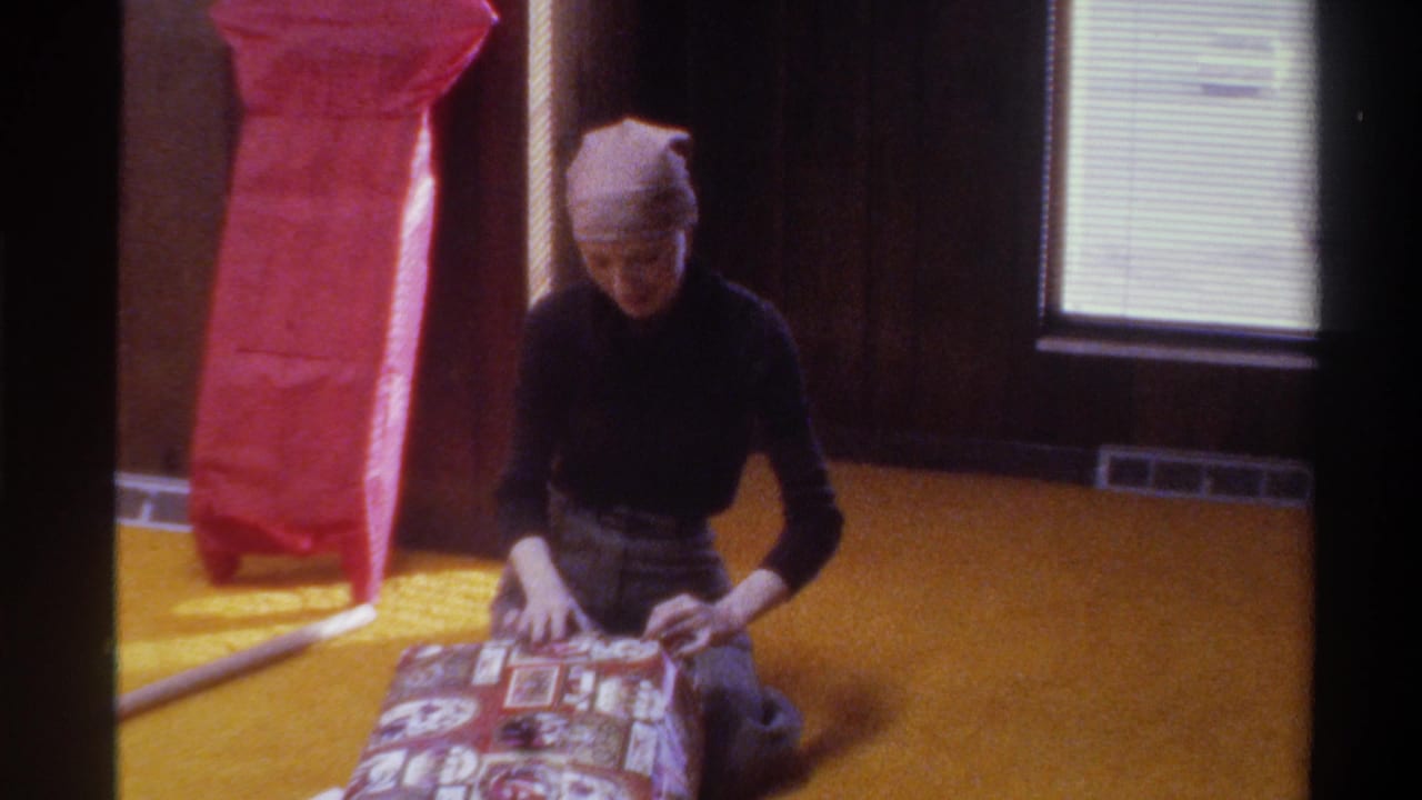 Woman in black turtleneck and knit beanie wraps gifts on yellow carpet in wood-paneled room, 8mm home movie, Gainesville Florida, circa 1975