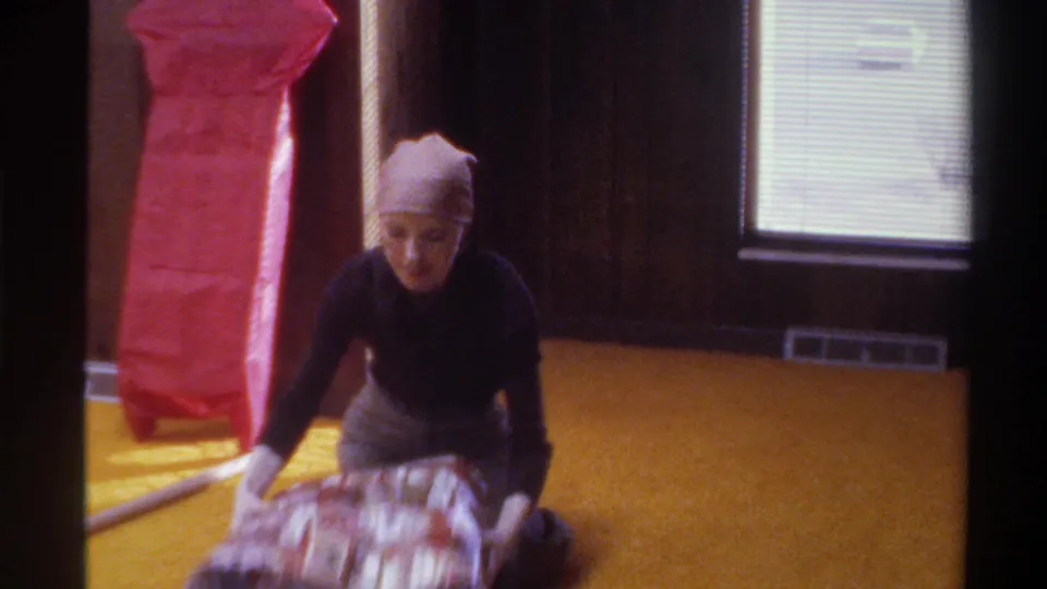 Woman leans forward over gift wrap spread on yellow carpet in wood-paneled room, 8mm home movie, Gainesville Florida, circa 1975