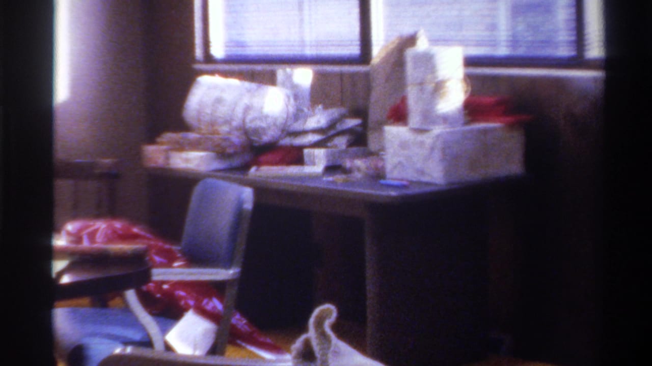Wide interior shot from a 1975 Gainesville, Florida home movie: Christmas gifts piled on dark table, blue chair at left, wood-paneled walls, horizontal blinds.