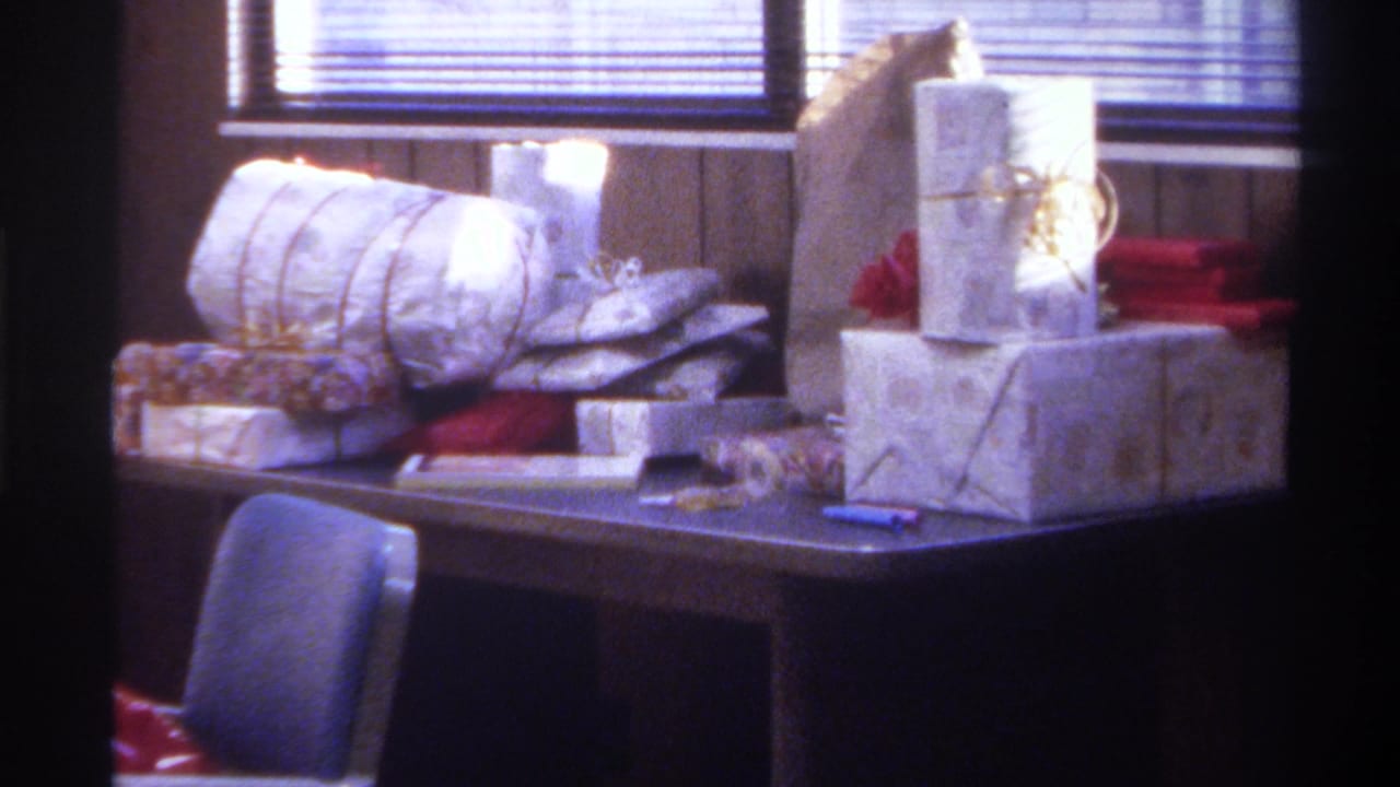 Room view with Christmas gifts on dark table, blue upholstered chair centered, red wrapping visible at right, wood-paneled walls and window blinds.