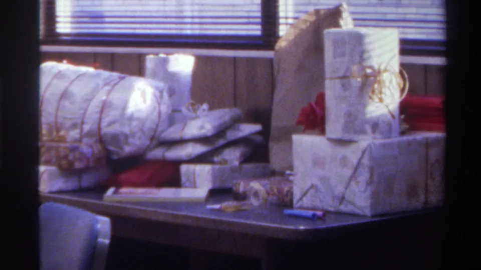 Close-up from a 1975 Gainesville, Florida home movie: cylindrical tube-wrapped gift and stacked silver-paper boxes with gold ribbon on a dark table, blue pen visible.