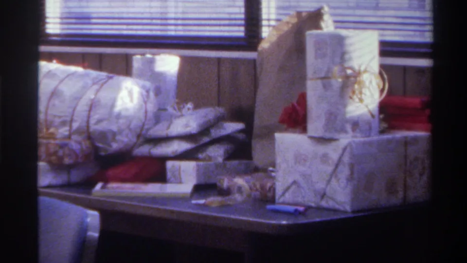 Close-up from a 1975 Gainesville, Florida home movie: stacked silver-wrapped Christmas gifts with gold ribbon detail, red items in background, wood-panel wall at right.