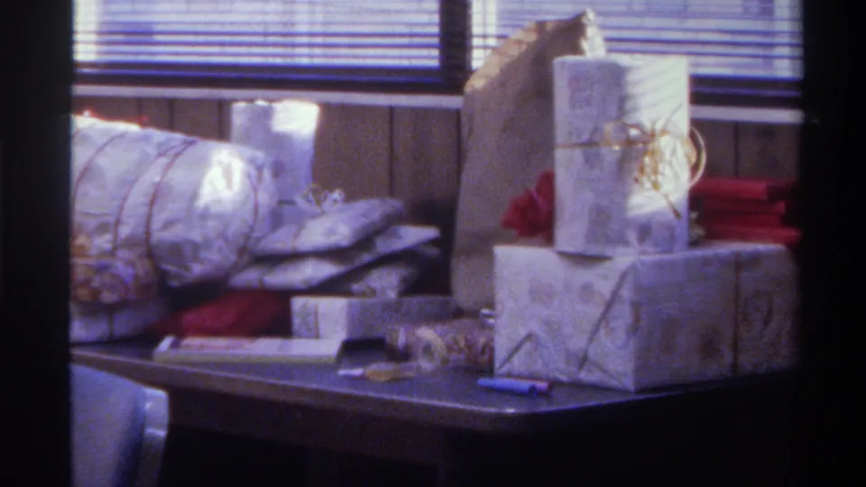 Wrapped gifts piled on a dark table with horizontal venetian blinds in background; Gainesville, Florida, 1975, 8mm home movie.
