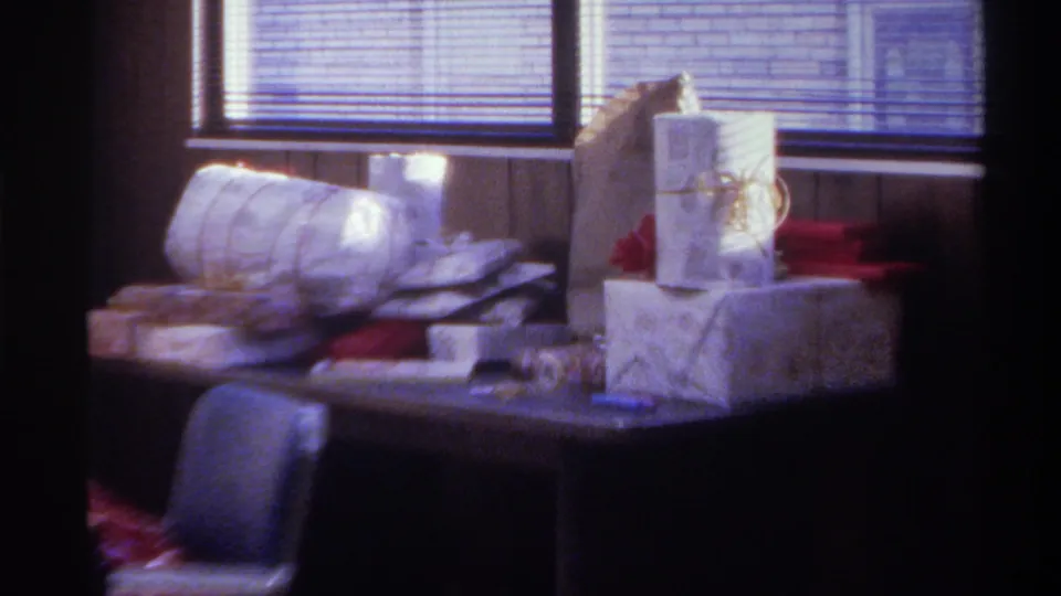 Wrapped gift pile on table with grey chair in foreground and backlit window blinds; Gainesville, Florida, 1975, 8mm home movie.