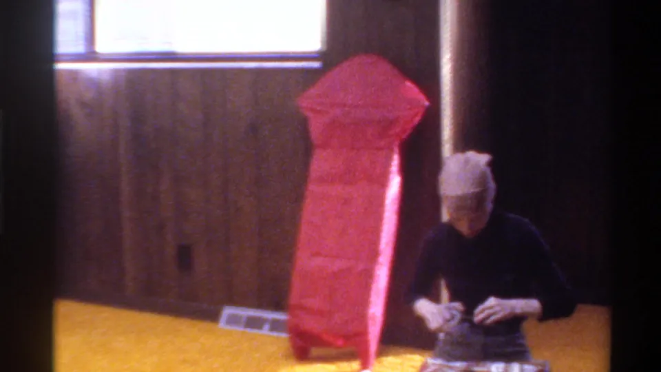 A woman preparing to wrap Christmas gifts beside a tall red-wrapped present in a wood-paneled interior, Gainesville, Florida, circa 1975.