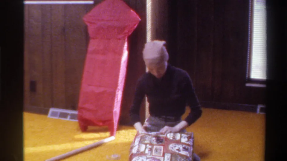 A woman wrapping a gift with patterned paper on gold carpet in a wood-paneled 1970s home, Gainesville, Florida, circa 1975, large red present nearby.