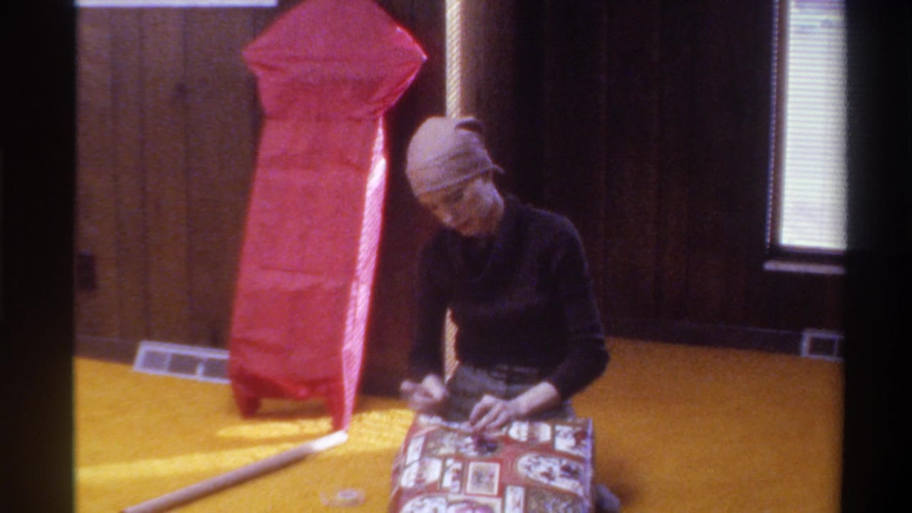 Woman in head wrap wrapping a patterned gift on yellow shag carpet; large red-paper-wrapped object behind her; 1975 American home interior.