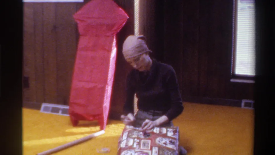 Woman in head wrap wrapping a patterned gift on yellow shag carpet; large red-paper-wrapped object behind her; 1975 American home interior.