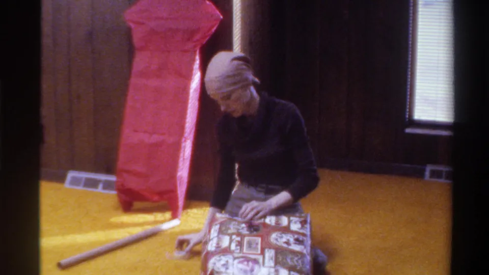 Woman in light head wrap continues wrapping a patterned gift on yellow shag carpet; large red-wrapped tall object at left; 1975 home interior.