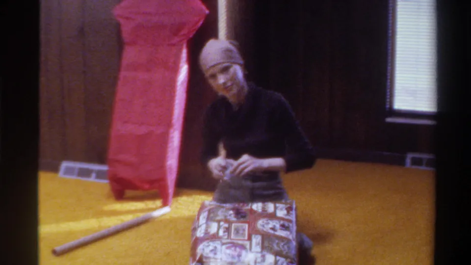 Woman pauses gift wrapping to look up, half-smiling; red-paper-wrapped tall object at left; yellow shag carpet and wood paneling; 1975 Florida home.